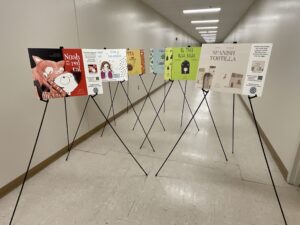 Six of the eight bilingual book walk covers are pictures here in a hall with indoor stands being used. The indoor stands are collapsible aluminum stands with adjustable viewing heights