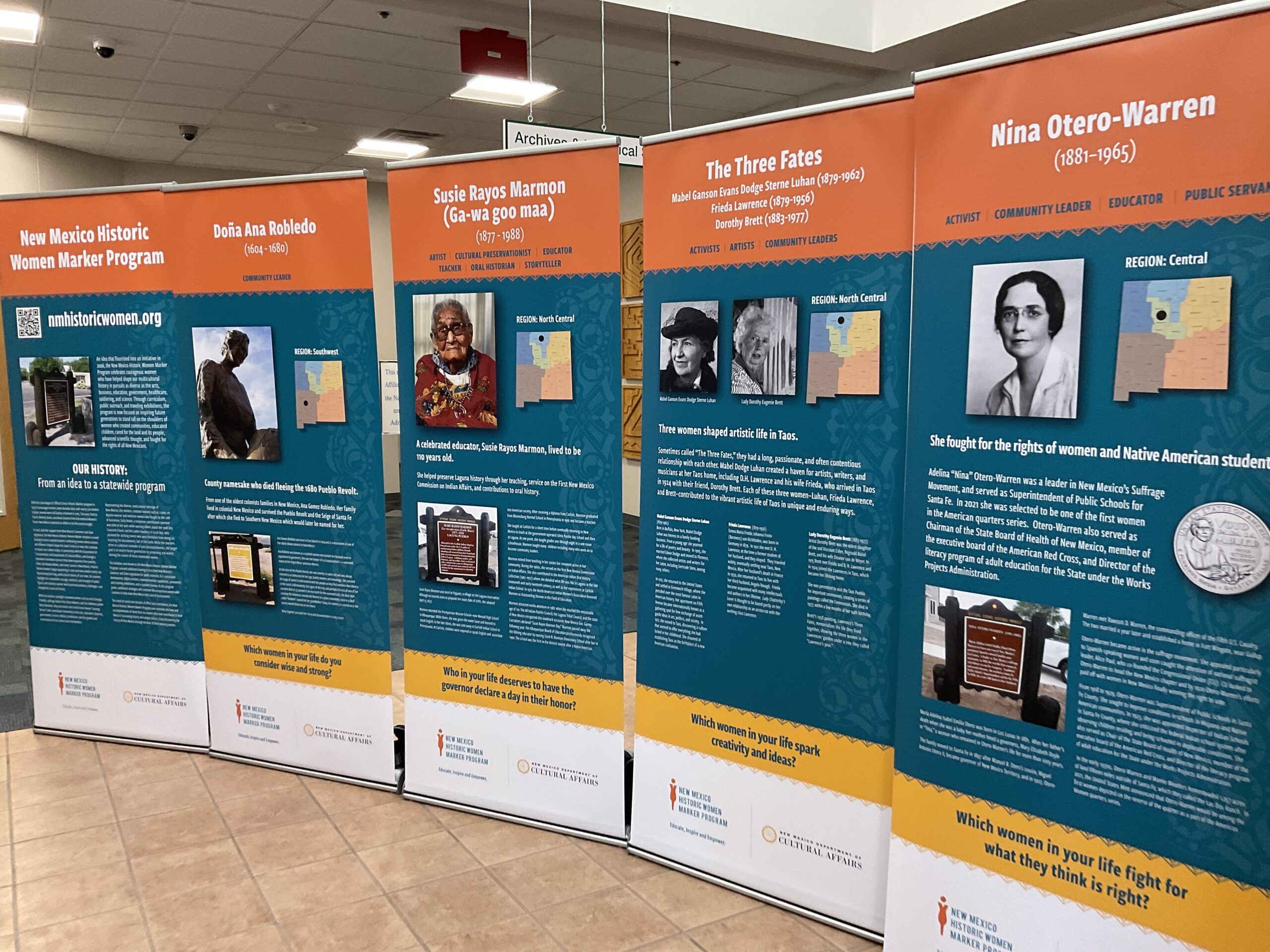 Five of the women's history banners displayed side by side. Titles from left to right: New Mexico Historic Women Marker Program, Dona ana Robledo, Susie Rayos Marmon, The Three Fates, and Nina Otero-Warren. The banners all display a picture and text. Banners of the historic women include their year of birth and death and a map locating where they are from in New Mexico.