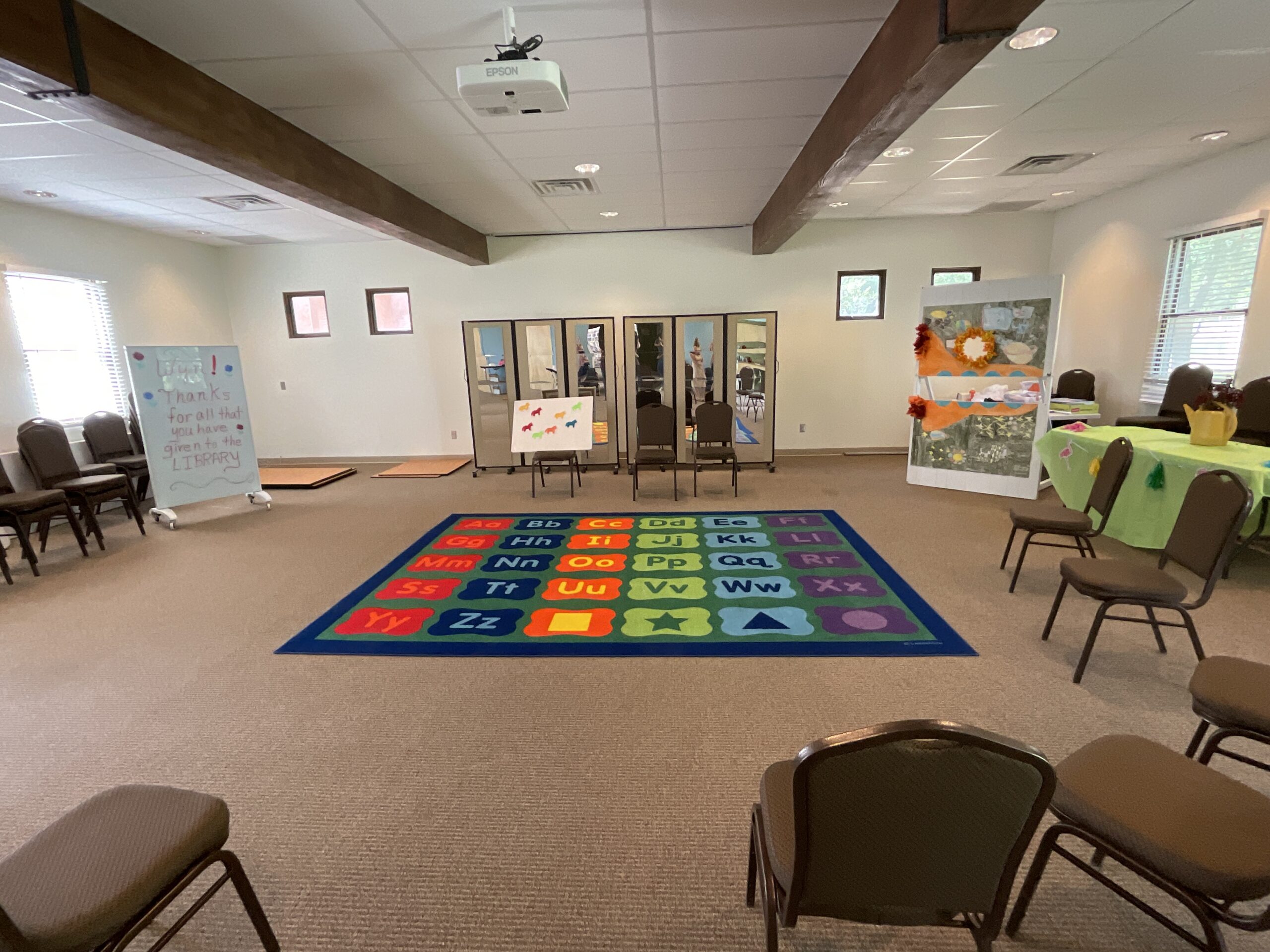 The indoor programming space of Bosque Farms Library is pictured. In the middle of the floor is an alphabet rug and around the parameter of the room are chair, tap mats, mirror, a photo booth prop, and a table. 