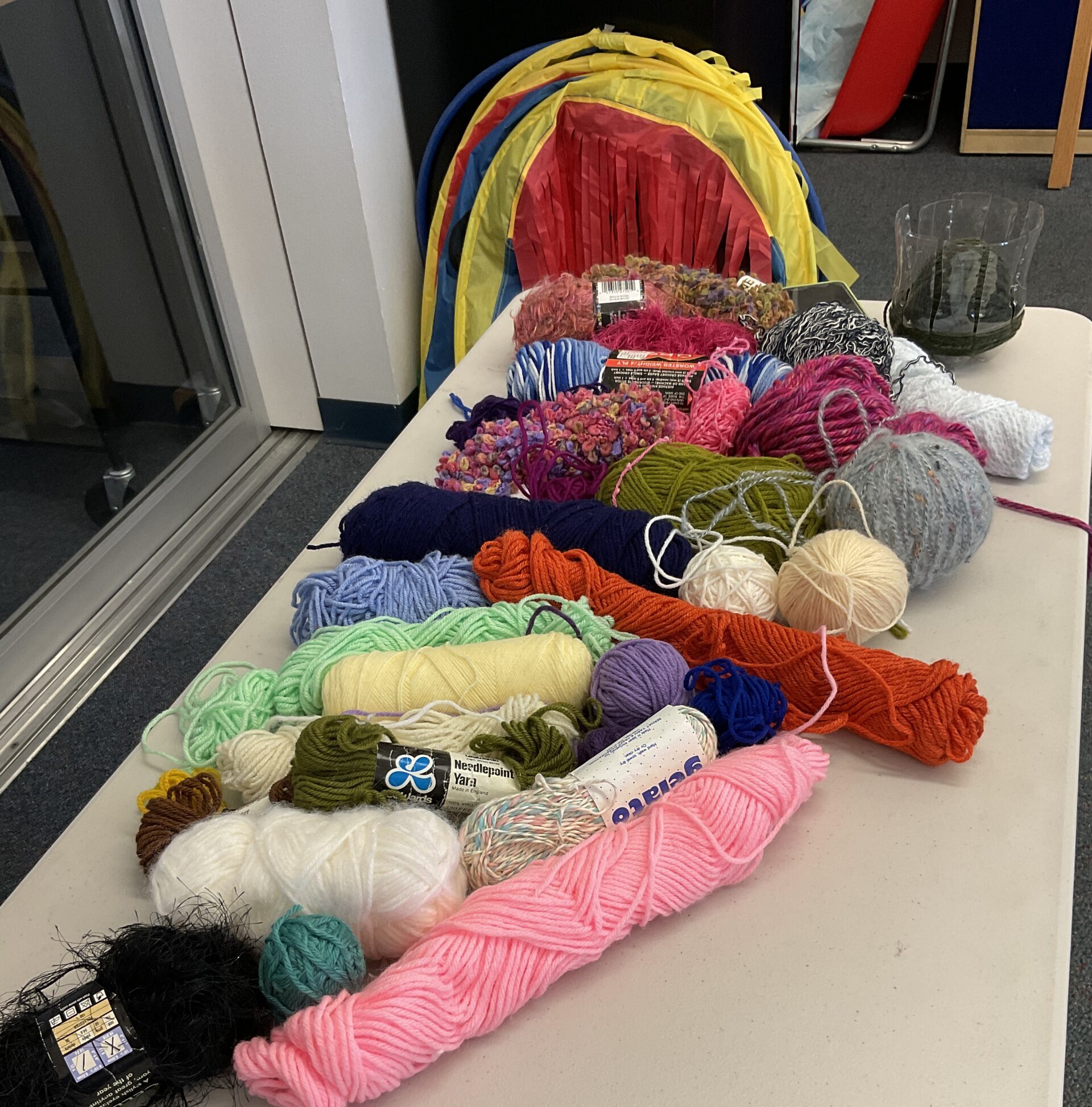 A white table with a large variety of yarn on top. There's a half-done example of a woven bowl on the far side of the table. 