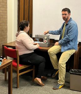 Woman sitting in front of sewing machine next to instructor who points to machine